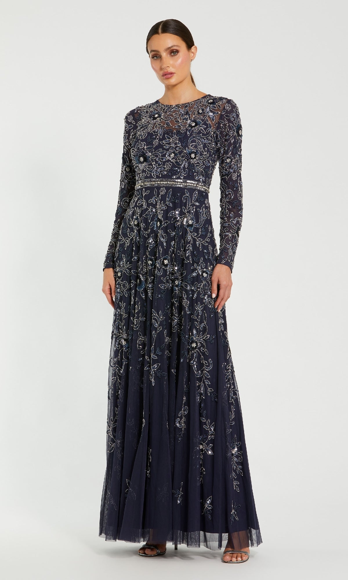 A woman wears the Mac Duggal 10093 Long Sleeve Beaded Formal Dress, a floor-length evening gown with silver sequin and bead embellishments, posing against a plain white background.